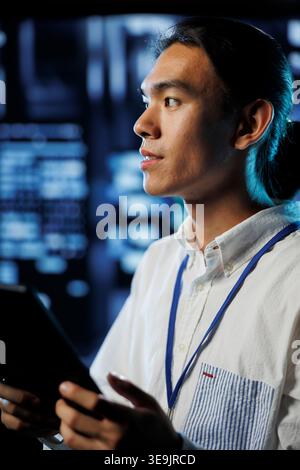 Developer in high tech facility uses tablet to prevent system overload during peak traffic periods. Asian programmer between server rows ensuring enough network bandwidth for smooth operations Stock Photo