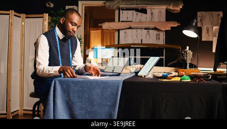 Professional couturier reviewing sketch, working with laptop and drawings to compare modern designs in atelier. Young tailor analyzing clothes models, needlework. Handheld shot. Stock Photo