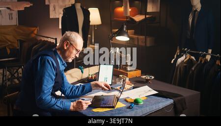 Senior craftsman shopping fabric online, using credit card at laptop in atelier. Experienced fashion designer buying material to create luxury clothing items, tailoring concept. Handheld shot. Stock Photo