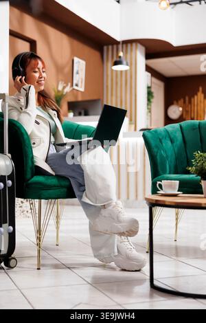 Happy Asian visitor waiting to reserve room while sitting at laptop with headphones on in contemporary hotel lobby. New guest lounging in resort foyer before booking room Stock Photo