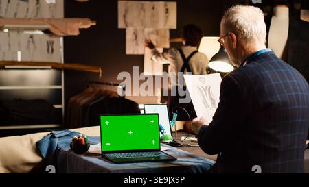 Senior designer with greenscreen analyzing sketches to create clothes for new fashion collection, tailoring concept. Experienced tailor working with chroma key isolated display. Stock Photo