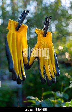 Yellow garden gloves tools dry on rope on greenery outdoor background during sunset. Spring summer gardening season. Concept of hand care hygiene and protection. Copy space Stock Photo