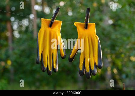 Yellow garden gloves tools dry on rope on greenery outdoor background during sunset. Spring summer gardening season. Concept of hand care hygiene and protection. Copy space Stock Photo