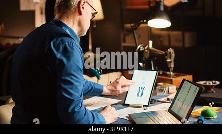 Male designer using laptop and tablet to check items on sketches, creating custom made clothes. Senior couturier at sewing table browsing device, creating fashion collection. Handheld shot. Stock Photo