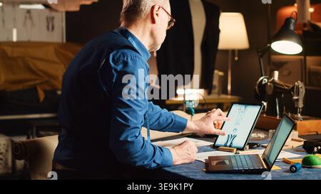 Old tailor works on clothes collection, comparing modern designs of tailored suit on laptop and tablet. Experienced fashion designer looking at sketches for new clothing line, craftsman job. Stock Photo