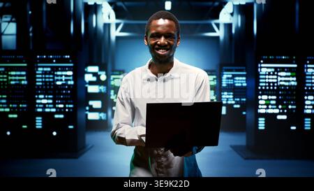 Computer scientist walking between data storage devices in computing server room, ensuring proper operations conditions. Serviceman monitoring high tech facility infrastructure energy consumption Stock Photo