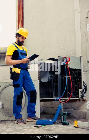 Experienced technician commissioned for outdoor air conditioner routine maintenance, looking for cooling system freon leaks. Skilled engineer doing hvac unit safety check, writing report on clipboard Stock Photo