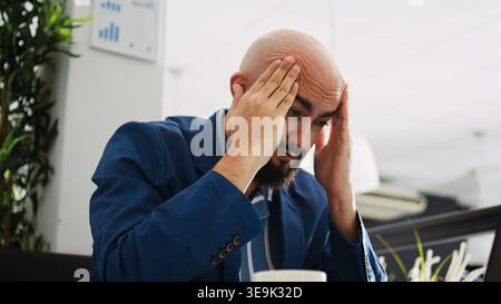 Burnout in coworking space. Businessman being tired and having migraine at work, trying to develop new ideas for startup improvement. Unwell overworked employee dealing with headache. Tripod shot. Stock Photo