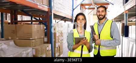 Two smiling Latin warehouse workers standing in stockroom aisle, banner Stock Photo