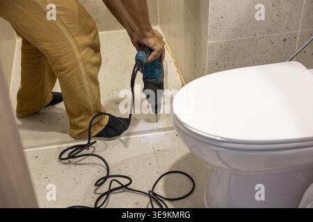 Focused man using power drill on floor tile in bathroom during home renovation project near white toilet, showing diligent repair work for interior re Stock Photo