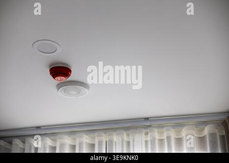 Red fire alarm smoke detector and white recessed led downlight fixture installed on residential ceiling providing reassuring safety and modern illumin Stock Photo