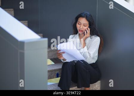 Young businesswoman sitting on office stairs, talking on smartphone and reviewing printed documents. Professional female employee managing reports, co Stock Photo