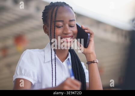 Close-up portrait of young Black business woman on a smartphone call, smiling while taking notes. Friendly communication, customer support, remote wor Stock Photo