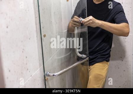 Focused adult man worker careful to install clear transparent glass shower door divider inside home bathroom renovation project holding metal handle f Stock Photo