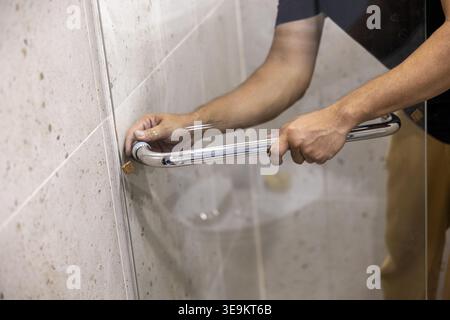 Careful worker installs metal handle on glass shower divider during bathroom renovation project showing focused expression while handling construction Stock Photo