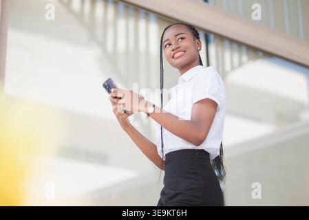 Low angle portrait of young Black business woman holding smartphone and looking away outdoors. Mobile connection, career confidence, online communicat Stock Photo