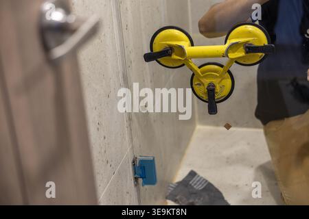 Focused worker install glass shower room divider using yellow suction cup tool inside tiled bathroom. Diligent man working carefully on home renovatio Stock Photo