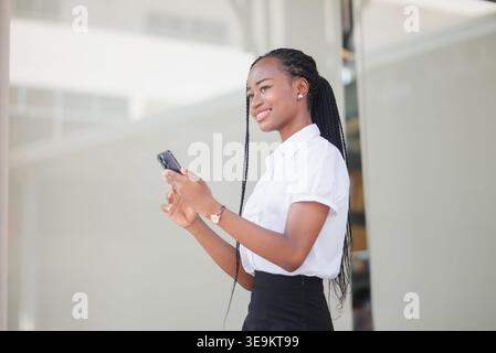 Side view of young Black business woman using smartphone outside an office building. Mobile communication, urban lifestyle, remote work, online messag Stock Photo