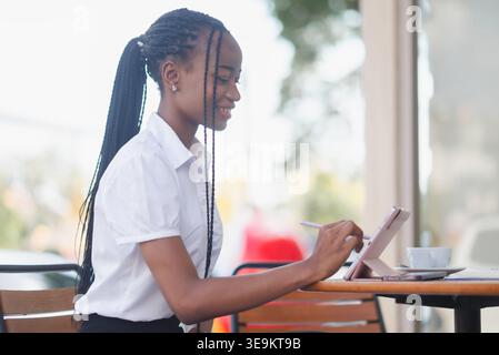 Side view of young Black business woman working on a tablet with stylus at a cafe table. Remote work, digital note taking, online planning, coffee bre Stock Photo