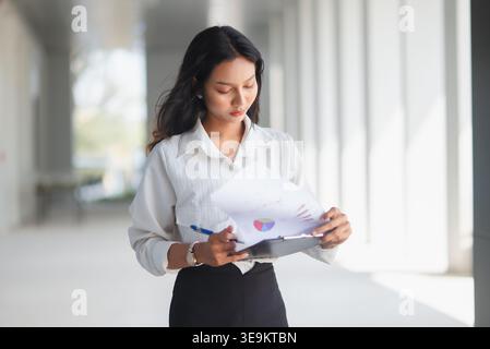 Young businesswoman reviewing financial report on clipboard in a bright modern office corridor. Professional female employee analyzing charts and pape Stock Photo