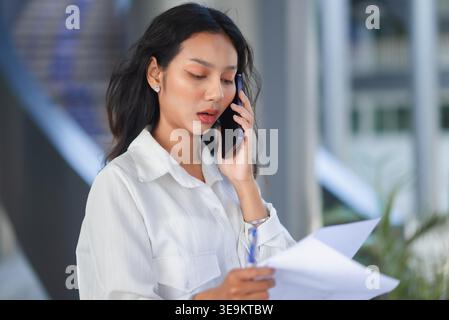 Young businesswoman talking on smartphone while checking paperwork in a modern office building. Professional female employee reviewing documents, taki Stock Photo