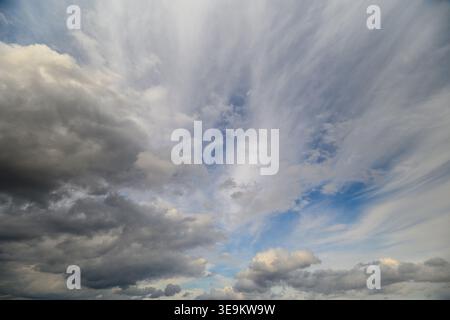 Dramatic layered clouds sweeping across sky with textured cirrus and heavy cumulus, sunrays piercing through breaks, looming stormfront over distant h Stock Photo