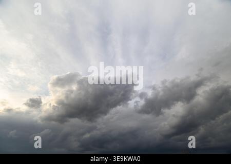 Dark storm clouds gather over horizon, dramatic sunrays piercing through breaks, heavy layered cumulonimbus suggesting approaching thunderstorm, moody Stock Photo