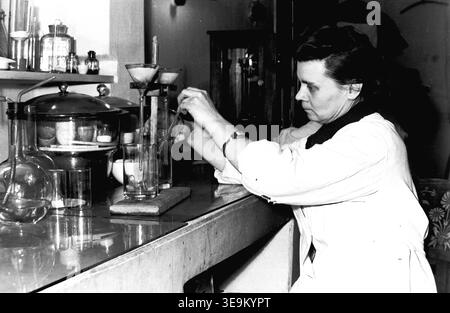 Slaviansk, 1950s, AIZ factory (Armature and Insulator Plant). A dedicated female Soviet laboratory technician wearing a white medical coat meticulously conducts chemical analyses using specialized glass beakers, funnels, and precision equipment. Seated at her organized testing bench, she ensures the strict quality control of raw materials for ceramic production, perfectly illustrating the critical scientific research and advanced technical standards of industrial manufacturing in the post-war era in peaceful Donbas. Stock Photo