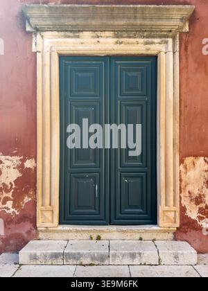 Green wooden double door framed by ornate stonework on a weathered red plaster wall, in Corfu, Ionian Islands, Greece. Stock Photo
