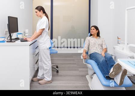 A dentist is entering patient data on a computer while a patient sits in a dental chair. The scene takes place in a modern dental office. This moment Stock Photo