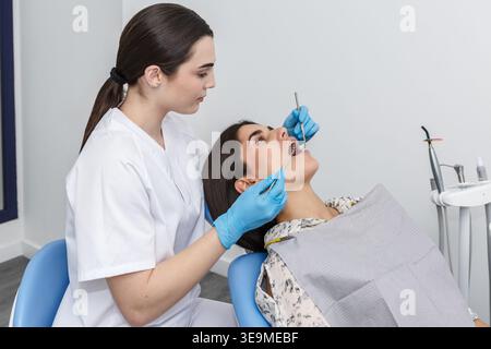 A dentist is examining a patient's teeth in a dental clinic. The patient is sitting in the dental chair, while the dentist uses tools for the check-up Stock Photo