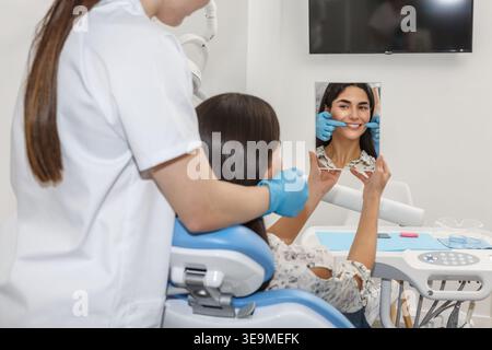 A dentist in a dental clinic is holding a mirror for a patient to see her smile. This scene takes place during a dental check-up session in a modern o Stock Photo