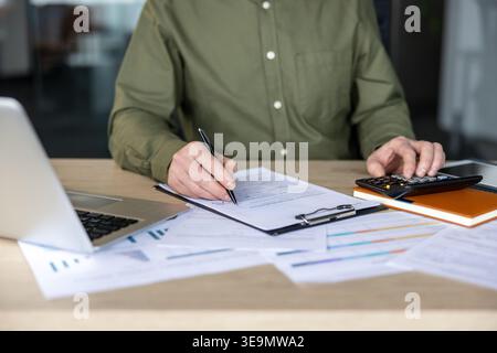 Accounting professional sitting at a desk and reviewing financial documents, signing paperwork, and using a calculator while working on a laptop in an office setting Stock Photo