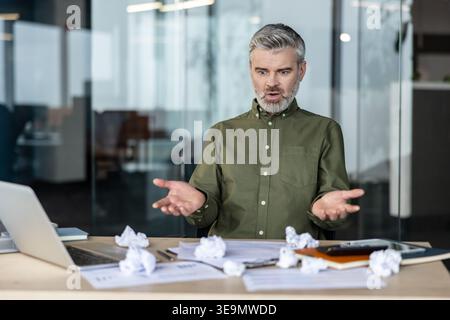 Frustrated man with gray hair expressing confusion and stress at a messy office desk, surrounded by crumpled paper, having problems with a business project Stock Photo