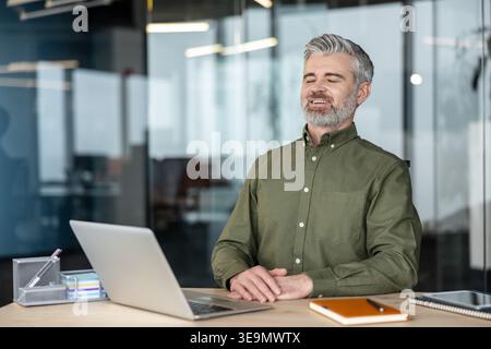 Peaceful businessman enjoying a moment of relaxation and smiling with closed eyes while sitting at his office desk with a laptop, finding balance and stress relief during the workday Stock Photo