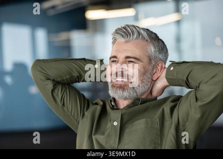 Smiling mature businessman with gray hair and beard leaning back, hands behind his head, enjoying a moment of relaxation and success in an office setting Stock Photo