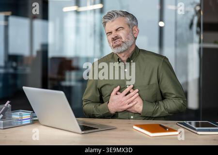 Mature man having a heart attack or experiencing severe chest discomfort while working in a modern office, showing symptoms of a health emergency caused by stress or illness Stock Photo