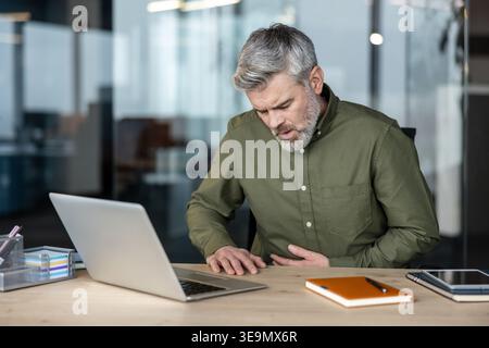 Mature man feeling sharp stomach pain while working on a laptop at his office desk, symbolizing workplace stress, illness, and health issues impacting productivity Stock Photo
