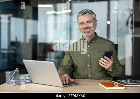 Mature businessman with grey hair and beard smiling for the camera while working at his modern office desk, typing on a laptop and holding a smartphone Stock Photo