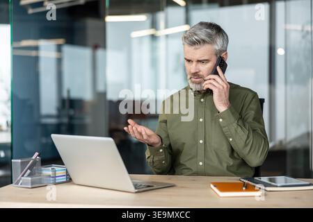Concerned man speaking on a mobile phone looking at his laptop screen, dealing with a problem or challenge while working in a modern office environment Stock Photo