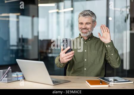 Mature man happily communicating via a virtual meeting or video call on his mobile phone in a modern office setup, actively engaging with a greeting gesture Stock Photo