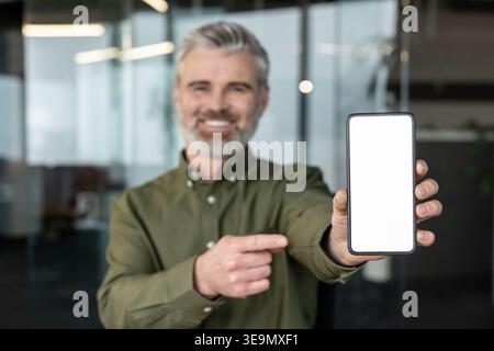 Happy mature businessman smiling and pointing at a smartphone with a blank screen, presenting an app or service in a modern office copy space for marketing and mockup use Stock Photo