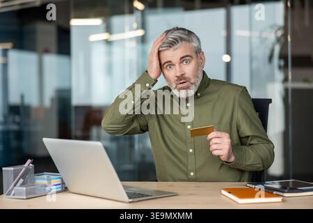 Mature man with a surprised and shocked expression touching his head, finding himself in an unexpected financial situation while holding a credit card and looking at a laptop in an office Stock Photo
