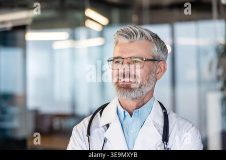 Mature male doctor wearing a white lab coat and stethoscope, happily looking away in a modern medical clinic, representing healthcare expertise, care, and professional medical services Stock Photo