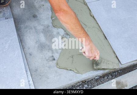 Worker spreading tile adhesive on a concrete base before installing outdoor ceramic tiles, close-up of construction process. Stock Photo