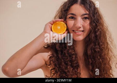 Woman with curly hair joyfully holds orange slice beside her face, smiling against a soft beige backdrop, representing freshness and vitality in lifestyle imagery Stock Photo