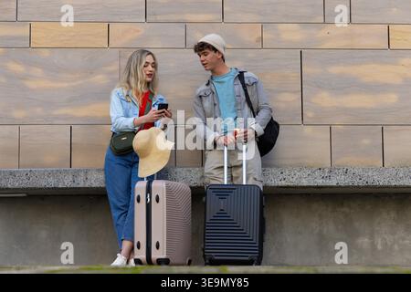 Couple standing at plaza in casual clothes, comparing smartphone next to suitcases and straw hat. Luggage, phone, travelers, backpack, sandstone, urban, square Stock Photo