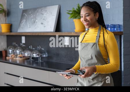 African American woman using tablet, reader at cafe counter wearing apron, mustard top, copy space. Staff, bakery, interaction, pastries, plants, interior, bright Stock Photo