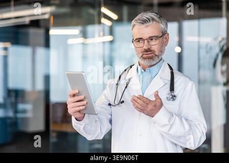 Mature doctor wearing a lab coat and stethoscope holding a digital tablet and gesturing, providing medical advice during an online consultation in a modern clinic setting Stock Photo