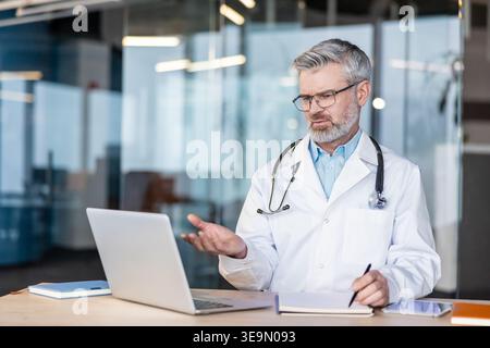 Experienced male doctor in a lab coat and stethoscope conducting a telemedicine video consultation on laptop from a modern medical office, explaining care remotely to a patient Stock Photo